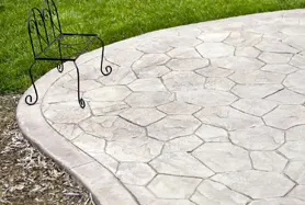 A stamped concrete patio with an irregular flagstone pattern in a light gray finish, bordered by a neatly curved edge separating the patio from a green lawn and mulched landscape bed. A decorative wrought-iron chair sits at the edge of the patio, showcasing the seamless integration of stamped decorative concrete with outdoor living spaces — representative of the stamped concrete and decorative finish services offered by Columbus Concrete Solutions.