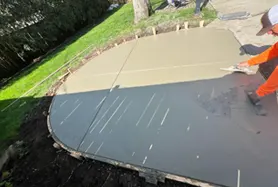 A concrete contractor in an orange safety vest uses a hand trowel to smooth and finish a freshly poured concrete patio or driveway section in a Columbus, Ohio residential yard, with the wet concrete surface showing a clean, even finish alongside an already-cured gray section. Green grass borders the curved edge of the pour, illustrating a professionally formed residential flatwork project typical of Columbus-area installations.