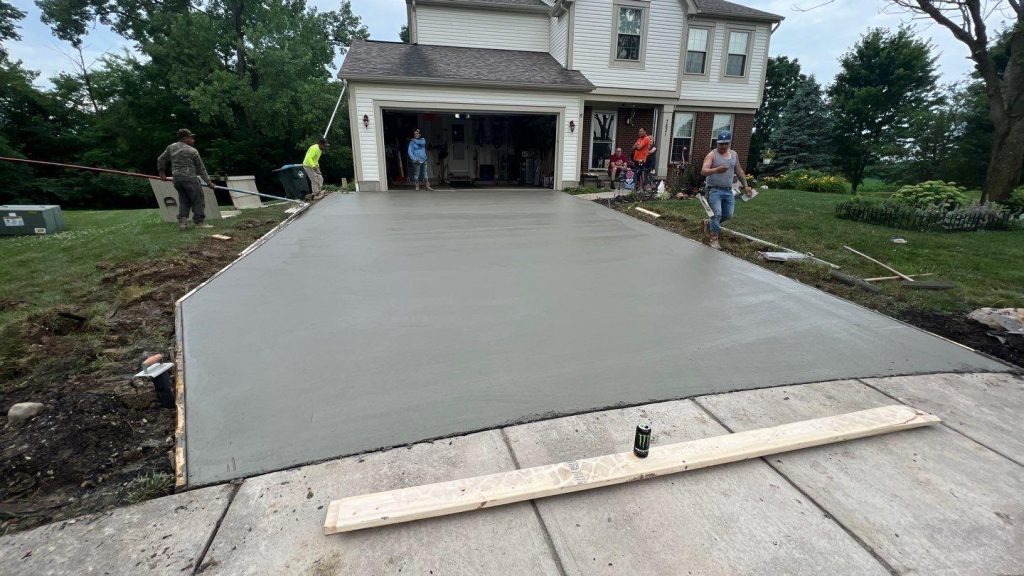 A freshly poured and smoothed concrete driveway leading to an open two-car garage of a two-story Columbus-area home, with a concrete crew finishing the surface using bull floats and hand tools while homeowners watch from the garage and front yard. The wet concrete shows a smooth, uniform finish bordered by wooden forms, with a straightedge board and a drink can resting at the foreground edge where the new pour meets the existing sidewalk. The lush green lawn, mature trees, and overcast sky reflect typical central Ohio summer conditions during a residential driveway replacement project.