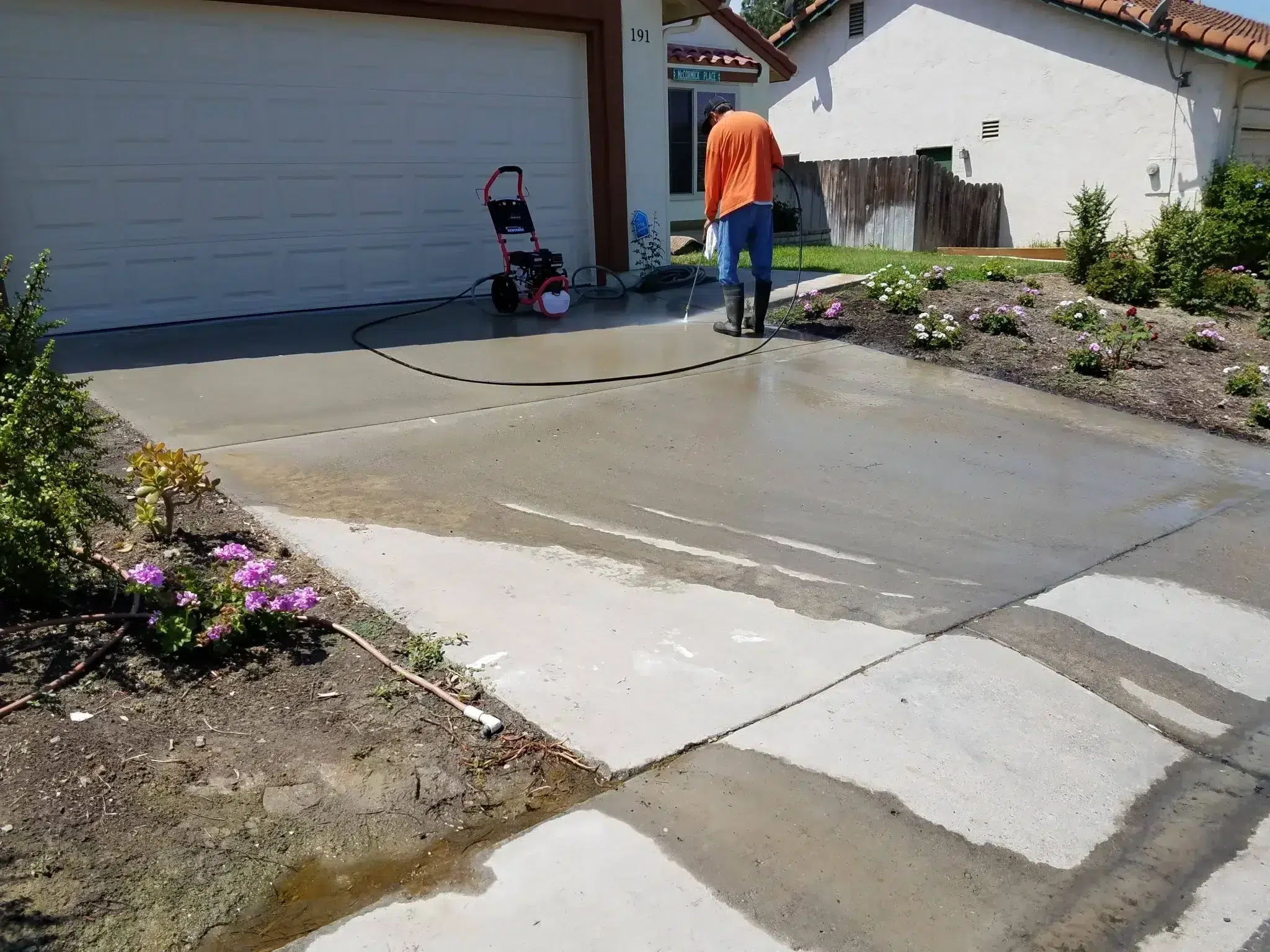Worker pressure washing a residential concrete driveway beside flowering landscape beds